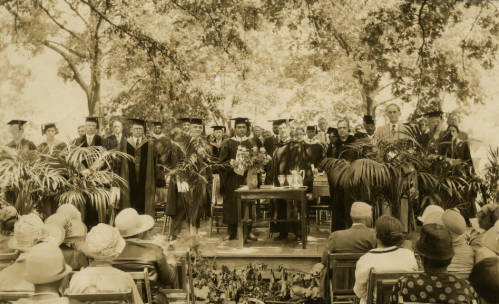 Approximately 30 people wearing caps and gowns on a stage in front of an audience. Dr. David Dallas Jones stands in the middle holding a piece of paper and looking at the camera with a serious look on his face. Black and white photograph.