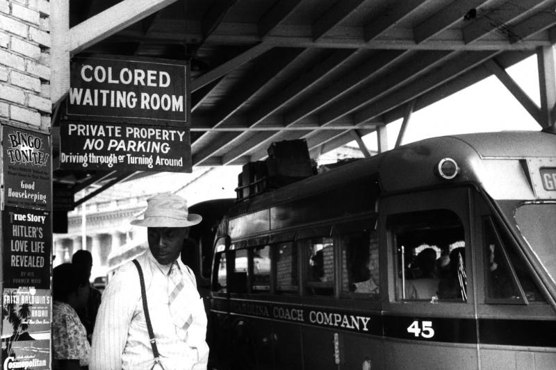 At the bus station A black man stands at a bus station beneath a sign reading "Colored Waiting Room." Black and white photo.