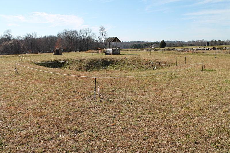 The Fort Dobbs site facing Fort Dobbs Road in Statesville, N.C. An open field with depressed craters. A small wooden well is in the background and a treeline marks the edge of the landscape.