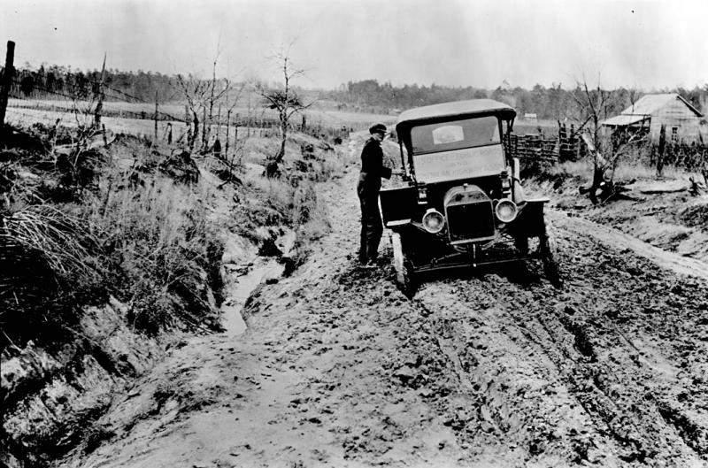 Automobile stuck in the mud A vintage automobile stuck in mud on a road. The road is cut through a hill, with banks on each side. A man is trying to get the car out of the mud. Their is a cabin in the background. Black and white.