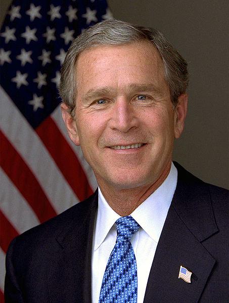 George W. Bush Photo of George W. Bush. He is smiling and standing in front of an American flag while wearing a suit.