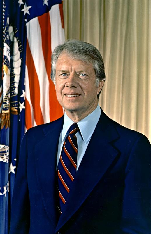 Jimmy Carter Picture of Jimmy Carter. He is wearing a blue suit, in front of yellow backdrop. There are American flags beside him. He has medium hair and no facial hair and is smiling.