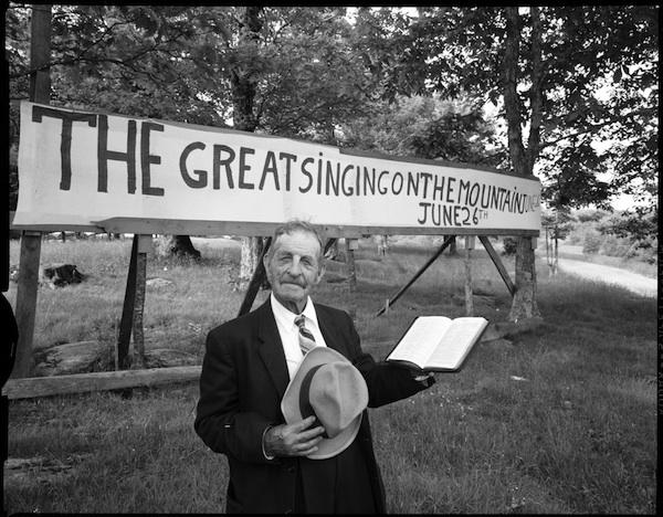 Joe L. Hartley Sr. in front of the Singing on the Mountain sign Joe L. Hartley Sr. in front of the Singing on the Mountain sign. He is holding his hat and a book. There are trees around.