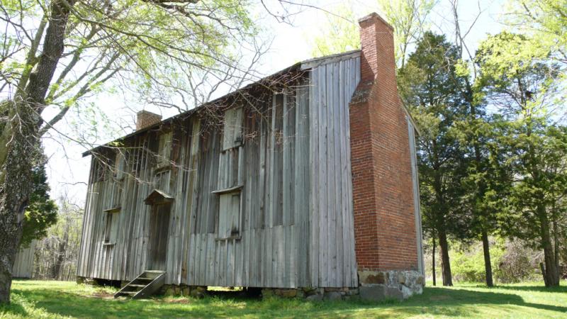 Exterior of slave house, Stagville Plantation A ramshackle wooden shack with a meager chimney sits against a woodland. It is sunny.
