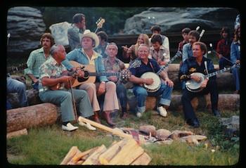 Roy Clark at Oral Roberts 1976 Television Special Five men surround a campfire and are playing instruments. There are people and large rocks in the background.