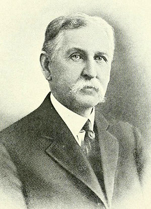 Black and white photo of Bellamy. He has medium graying hair, a thick moustache, and he is wearing a suit. 