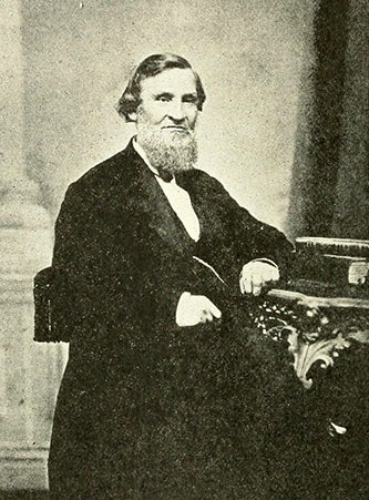 Cook seated for a photo. He has a prominent beard and medium hair. He is wearing a suit. 